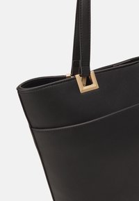 Czarna skórzana torba tote o wyraźnym kształcie, z kwadratowym złoconym akcentem i gładką teksturą. Prosty i funkcjonalny design.