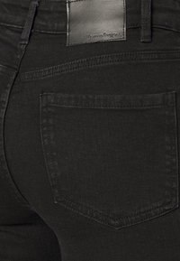 Jeans de ganga preta com um bolso traseiro com detalhes costurados e uma etiqueta de marca em couro na cintura, apresentando um acabamento mate.