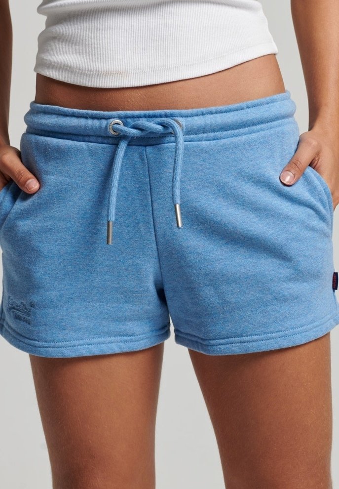 Personne portant un short bleu clair à cordon et un débardeur blanc côtelé, debout les mains dans les poches devant un fond uni.