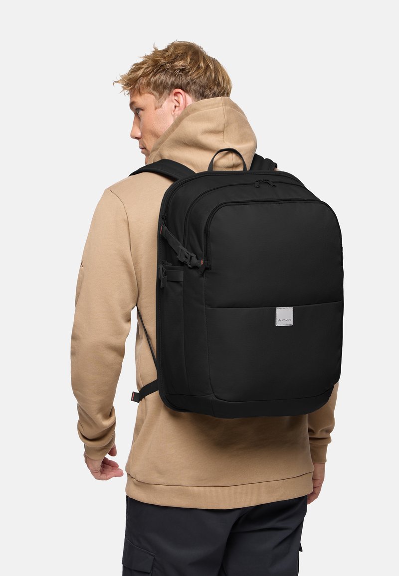 Joven con sudadera con capucha beige que lleva una mochila negra grande con varios compartimentos, de pie frente a un fondo claro y liso.
