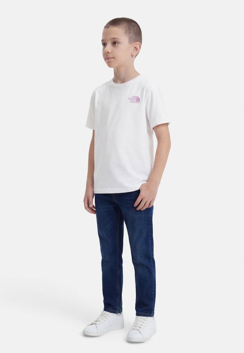 Chłopiec stojący w białej koszulce z krótkim rękawem z logo, ciemnoniebieskich dżinsach i białych tenisówkach na białym tle.