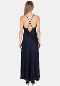 Donna con lunghi capelli biondi che indossa un abito lungo blu notte con spalline incrociate sottili e stivali neri con tacco, in piedi di spalle.