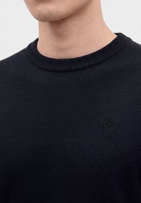 Homme portant un pull en maille noir à col rond avec un logo noir brodé subtil sur la poitrine, sur un fond uni.