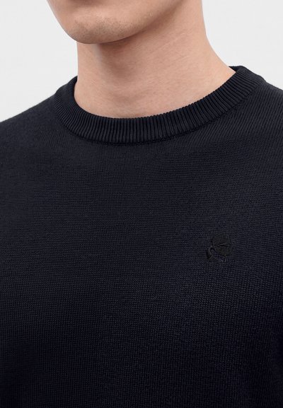 Homme portant un pull en maille noir à col rond avec un logo noir brodé subtil sur la poitrine, sur un fond uni.