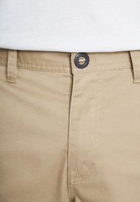 Close-up van de taille van beige broek met zwarte knoop en riemlussen, gedragen met een witte overhemd die is ingestopt.
