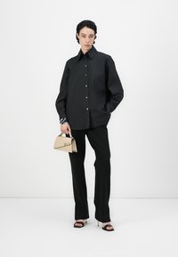 Camicia nera oversize con chiusura a bottoni, maniche lunghe e dettaglio logo bianco. Abbinata a pantaloni neri e una borsa beige. I tacchi completano l'outfit.