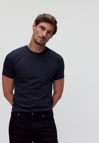 Marineblaue kurzärmlige T-Shirt in einem taillierten Design, aus glattem Baumwollstoff, kombiniert mit schwarzen Jeans und einem neutralen Hintergrund.
