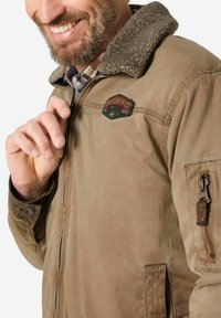Braune Canvasjacke mit einem flauschigen, braunen Kragen, einem Reißverschlussfach am Ärmel und einem gestickten Logo-Patch auf der Brust.