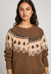 Maglione a maglia marrone con motivo geometrico in beige, arancione e teal. Presenta un collo rotondo e polsini a coste. Texture morbida.