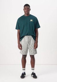 Groene katoenen T-shirt met gele branding, gecombineerd met lichtgrijze geblokte shorts, zwarte sneakers en zwart-wit gestreepte sokken.