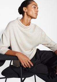 Maglione morbido di un beige chiaro, con collo a cascata e maniche a tre quarti. Modella seduta su una sedia nera, indossando pantaloni neri larghi.