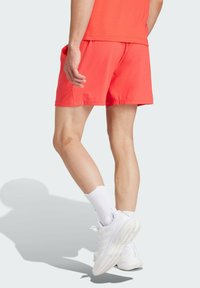 Red athletic shorts with an elastic waistband, smooth texture, and side pockets, paired with white athletic shoes and socks.