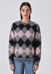 Donna che indossa un maglione a motivi argyle grigio, rosa e nero con maniche lunghe e jeans blu, in piedi di fronte a uno sfondo bianco uniforme.