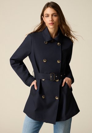 Frau mit langem braunem Haar, die einen marineblauen, zweireihigen Mantel mit Gürtel und großen Knöpfen sowie hellblaue Jeans trägt, mit den Händen in den Taschen.