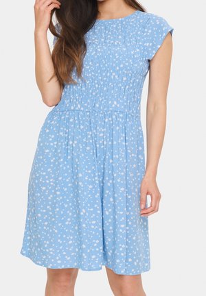 Hellblaues Kleid mit kurzen Ärmeln und gerafftem Oberteil, weißem Blumenmuster, das locker knapp über das Knie fällt. Glatte Stoffstruktur.