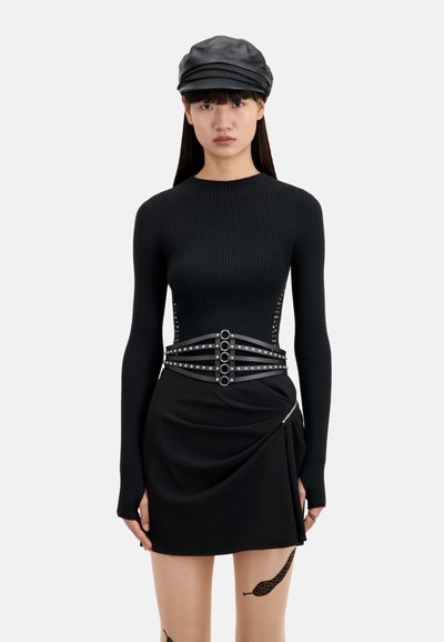 Jonge vrouw in een zwart ribbed langslavend shirt, zwarte leren pet, zwarte rok met asymmetrische drape, gestikte riem en een slangentatoeage op haar bovenbeen.