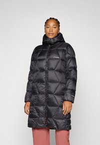 Osoba stoji u crnoj puf jakni s kapuljačom, dizajniranoj za toplinu, u kombinaciji sa svijetlim hlačama na neutralnoj pozadini.