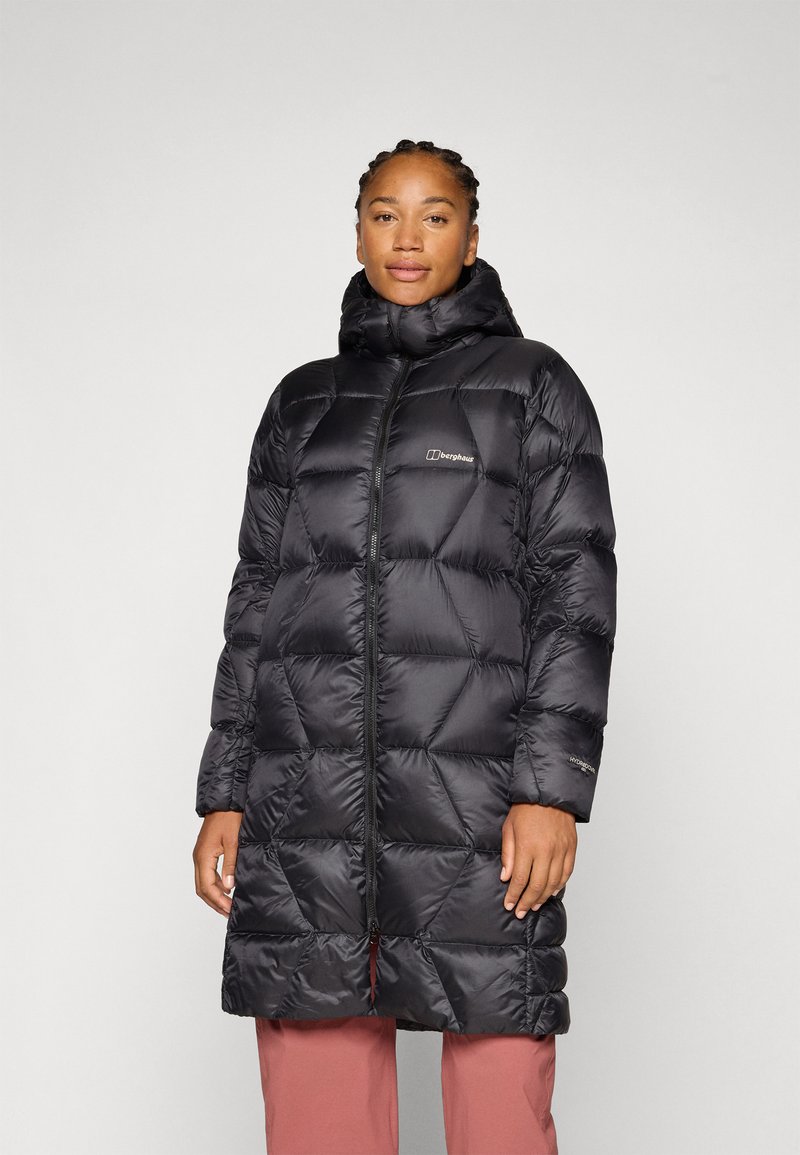 Osoba stoji u crnoj puf jakni s kapuljačom, dizajniranoj za toplinu, u kombinaciji sa svijetlim hlačama na neutralnoj pozadini.