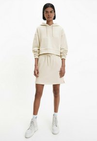 Sudadera recortada beige con cordón, combinada con una falda a la altura de la rodilla a juego que presenta aberturas laterales. Tejido texturizado y detalle bordado.