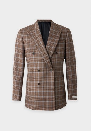 Homme portant un blazer croisé marron à carreaux, une chemise blanche, un pantalon gris foncé et des chaussures marron, debout devant un fond uni.