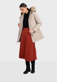 Béžová péřová bunda s umělou kožešinou na kapuci, přiléhavý střih, doplněná skládací červenou sukní a černými kozačkami ke kolenům.