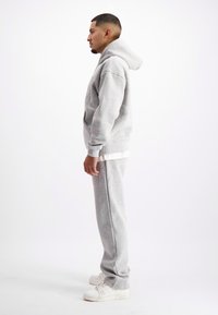 Man die zijwaarts staat en een lichtgrijze hoodie draagt, bijpassende trainingsbroek, witte sneakers en een wit shirt dat onder de hoodie uitsteekt.