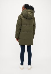 Manteau en duvet de couleur vert olive avec une capuche et un design matelassé, présentant des coutures horizontales et une longueur mi-cuisse. Porté sur un pantalon foncé et des baskets.