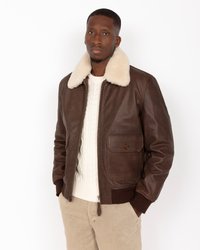 Chaqueta bomber de cuero marrón con cuello de borrego color crema, cierre de cremallera y bolsillos laterales. Llevada sobre un suéter de punto en trenzas color crema.