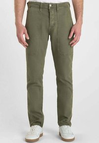Pantalons en coton vert olive avec une coupe droite, dotés de poches avant et de coutures renforcées, associés à des baskets blanches.