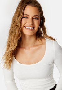 Witte geribbelde longsleeve top met een brede ronde hals. Het model heeft golvend haar en glimlacht, met natuurlijke make-up en gouden creolen.
