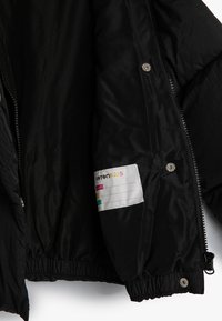 Schwarze Pufferjacke mit glatter Außenseite, elastischen Bündchen und Saum. Innen befindet sich ein Etikett mit bunten Markierungen für Namen. Mit Reißverschluss.