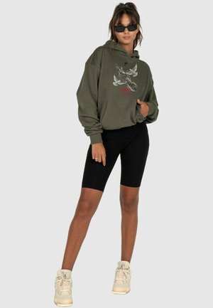 Felpa verde oliva con design grafico, tasca frontale e polsi a coste, abbinata a pantaloni biker neri e sneakers crema.