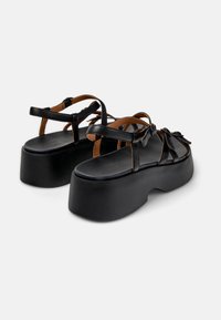 Černé platformové sandály s tenkými hnědými koženými pásky, mají texturovanou podrážku a zaoblený siluetu. Nastavitelné uzávěry.