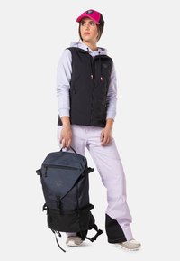 Schwarze isolierte Weste über einem helllila Hoodie, kombiniert mit weißen Hosen. Ein dunkelblauer und schwarzer Rucksack wird getragen, mit verstellbaren Trägern.