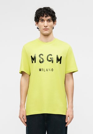 Kiharatukkainen mies, joka pukeutunut kirkkaan keltaiseen MSGM Milano -paidant ja tummiin housuihin, seisoo yksinkertaisen vaalean taustan edessä.