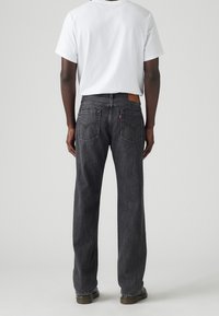 Muž oblečený v černých rovně střižených džínách a bílém tričku s krátkým rukávem, stojící zády k nám na jednobarevném světle světlém pozadí.