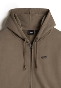 Brązowa bluza z zamkiem błyskawicznym, wykonana z teksturowanej tkaniny, z kapturem regulowanym sznurkiem oraz małym czarnym logo Vans na lewej piersi. Gładki, casualowy design.