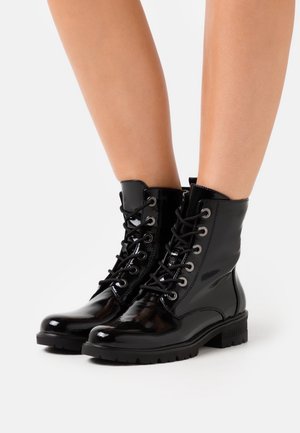 Glanzende zwarte veterlaarzen tot aan de enkel, gedragen op blote benen, met grove zolen en zilverkleurige veteroogjes, afgebeeld tegen een witte achtergrond.