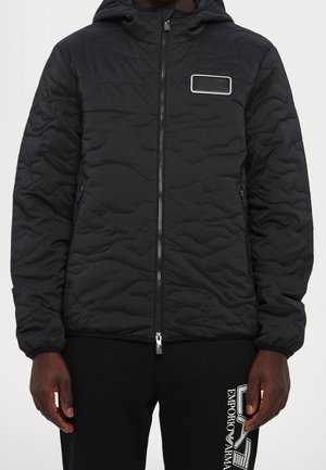 Personne portant une veste matelassée noire avec fermeture éclair devant et un patch sur la poitrine, associée à un pantalon noir avec un logo blanc Emporio Armani.