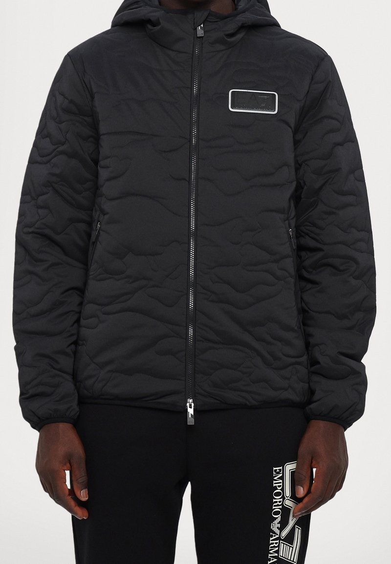 Personne portant une veste matelassée noire avec fermeture éclair devant et un patch sur la poitrine, associée à un pantalon noir avec un logo blanc Emporio Armani.
