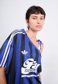 Camiseta de rayas negra y azul de gran tamaño con el logo de Adidas en blanco, un hombro al aire y tirantes finos en el lado expuesto.