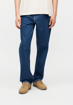 Person trägt lockere blaue Jeans und hellbraune Wildlederschuhe, steht vor einem schlichten weißen Hintergrund.