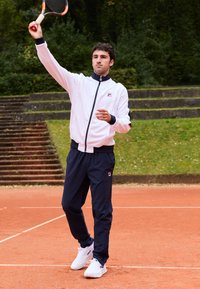 Casaco branco com fecho de correr e detalhes em azul-marinho, calças de desporto azul-marinho e ténis brancos; jogador a segurar uma raquete de ténis num court de terra batida.