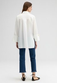 Witte langslavende shirt met een kraag en uitlopende manchetten, gecombineerd met blauwe jeans met uitgescheurde zoom en zwarte platte schoenen.