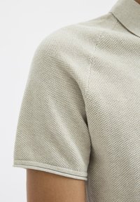 Lys grå polo skjorte med korte ermer, med et teksturert strikkemønster og en ribbestrikket krage. Detaljer inkluderer en kontrasterendekant på ermene.