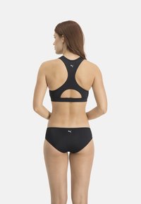 Schwarzer Sport-BH mit Racerback-Design, der einen Ausschnitt am Rücken und ein kleines Logo aufweist. Ergänzt durch passende schwarze Bikinihosen.