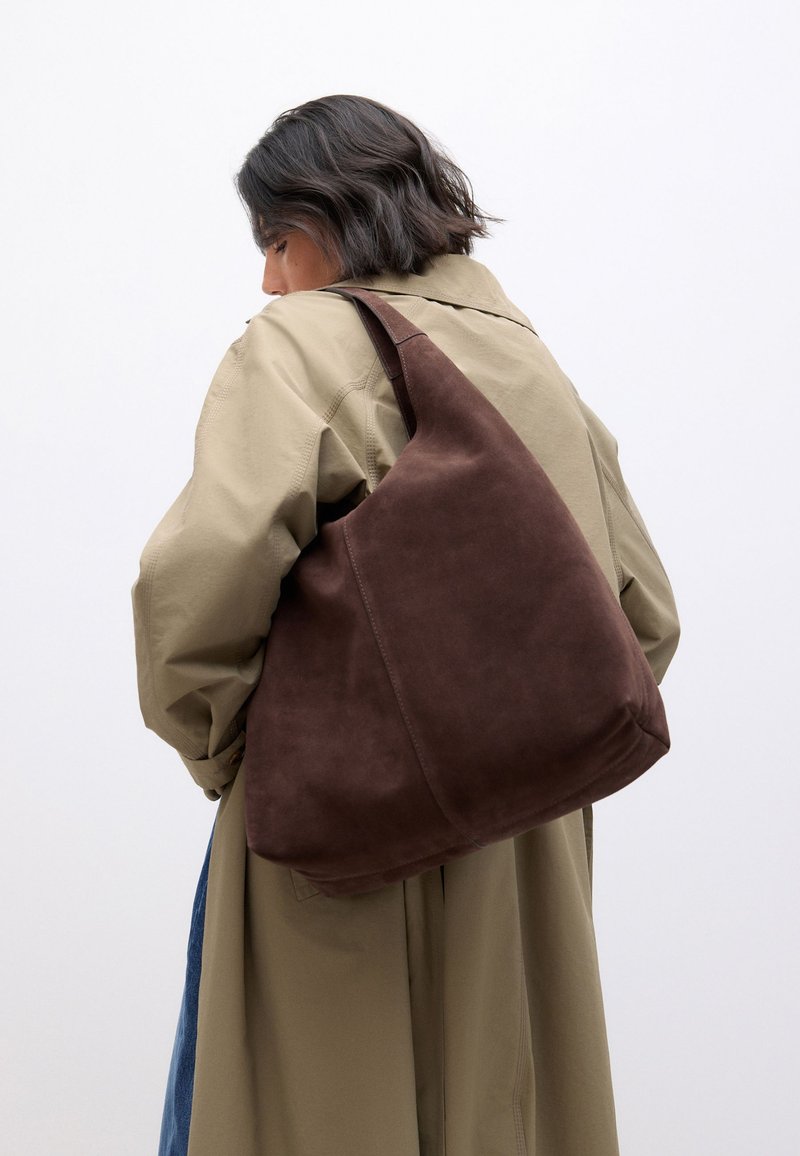 Femme portant un trench beige et un grand sac à bandoulière en daim marron sur l'épaule, sur un fond blanc uni.