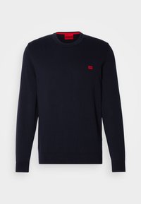 Granatowy sweter dzianinowy z długimi rękawami, okrągłym dekoltem i ściągaczami na rękawach. Posiada małą czerwoną naszywkę z logo na piersi. Gładka faktura.