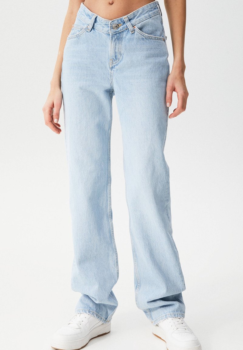 Jean en denim bleu clair avec une taille haute, une coupe droite et des détails de couture subtils. Porté avec des baskets blanches.