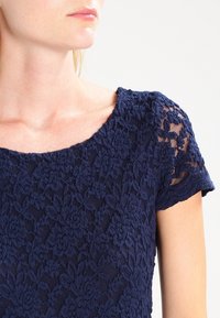 Námořnická modrá krajková blůza s krátkými rukávy, zdobená květinovými vzory a texturovanou tkaninou. Kulatý výstřih a mírně průsvitné detaily.